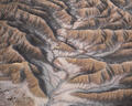 A winding desert wash carves through layered Utah badlands in soft gold and gray tones.
