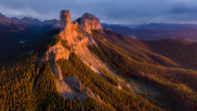 At sunset a tall, lone standing rock shines in the sun  and is surrounded by pine trees that flow down into the valley with gray clouds in the sky and mountain