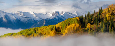 Bare jagged mountain tops interrupt the snow covered peaks in the background with lemon-lime colored aspen trees on the hillside in front and clouds hovering th