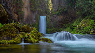 A two-layer waterfall with vertically gouged rock stretching up and lush green foliage and mossy covered rocks meeting the pool of water.