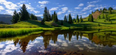 Lake in the mountains reflects surrounding spruce trees, grasses, mountains and sky with clouds.