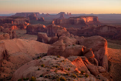 A southwest desert scene with mesas and buttes throughout the landscape of reds, pinks and tans at sunset with the sun spotlighting the scene from the left.