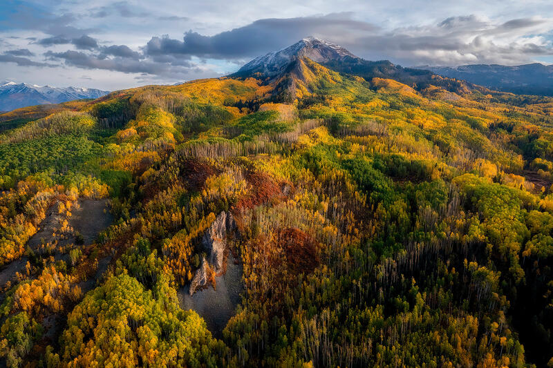 Mountain stands off in the distance surrounded by aspen trees in fall colors on hills descending from the mountain.
