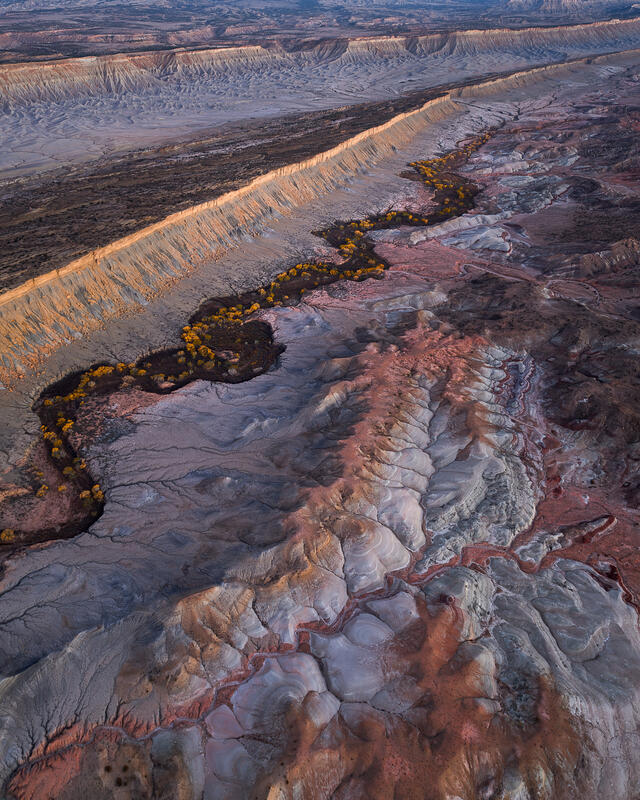 Winding river with golden cottonwoods through layered Utah badlands beneath a long escarpment.