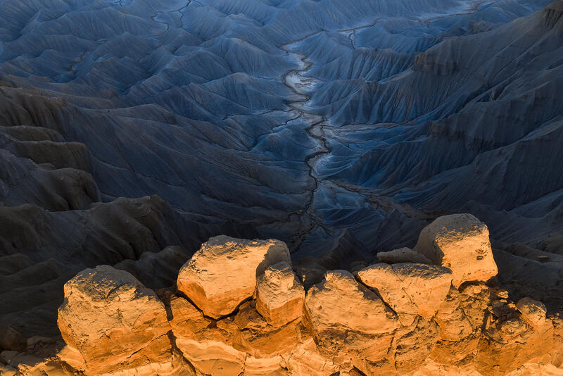 Golden light meets deep blue badlands as a winding channel leads through Utah’s desert below.