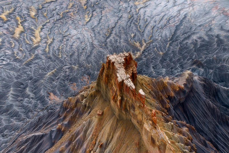 A solitary rock spire rises above intricate badlands patterns in Utah from an aerial perspective.