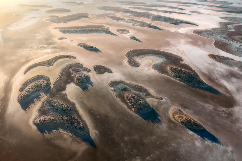 An aerial abstract of island-like formations and long shadows across an Australian dried lake bed.
