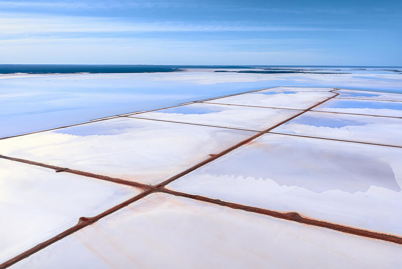 An aerial view of geometric salt ponds and reflective water under soft blue skies in Australia.