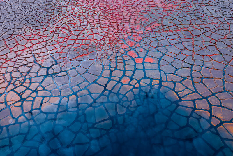 An abstract aerial of cracked mineral patterns blending vivid reds and cool blues across the Australian landscape.