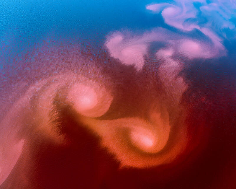 An abstract aerial composition of swirling mineral colors blending into fiery reds and cool blues across the Australian landscape.