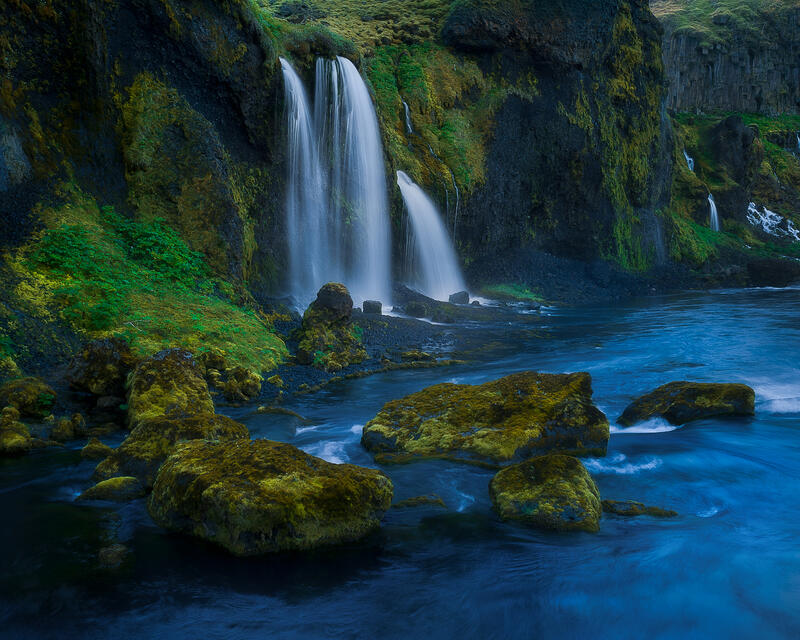 Hidden waterfalls descend through lush volcanic cliffs into a peaceful Icelandic river.
