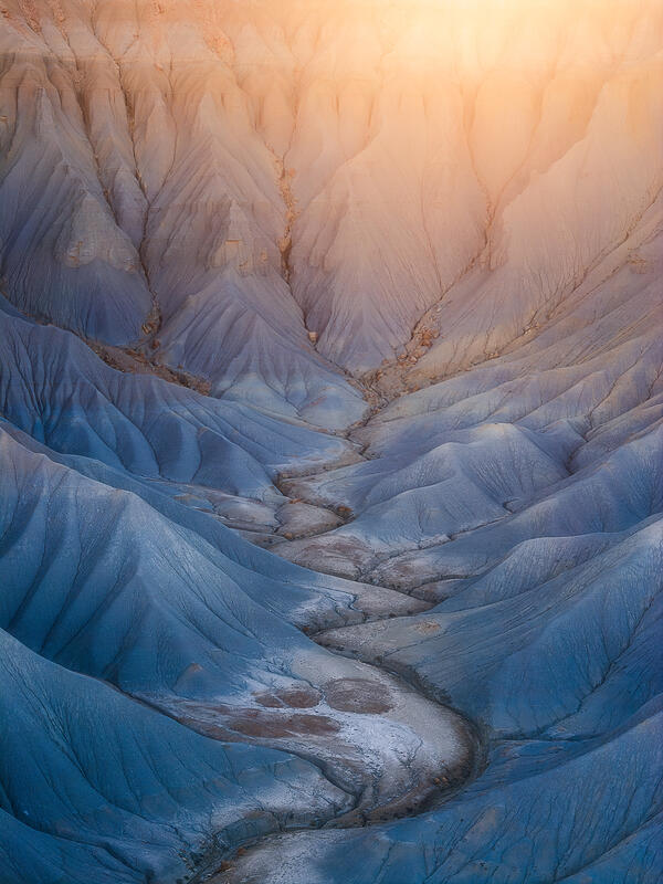 Blue badlands glow under soft sunrise light as a winding channel leads through Utah’s desert.