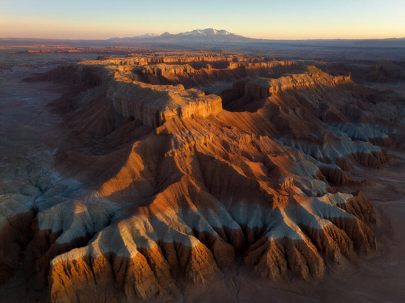 A sunlit Utah mesa rises like a fortress above sculpted badlands beneath distant mountain peaks.