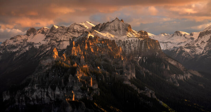 A mountain ridge is seen with snow on the top as the sun hits the top of the ridge at sunset and the spruce trees on the slopes are left in the shade in this dr