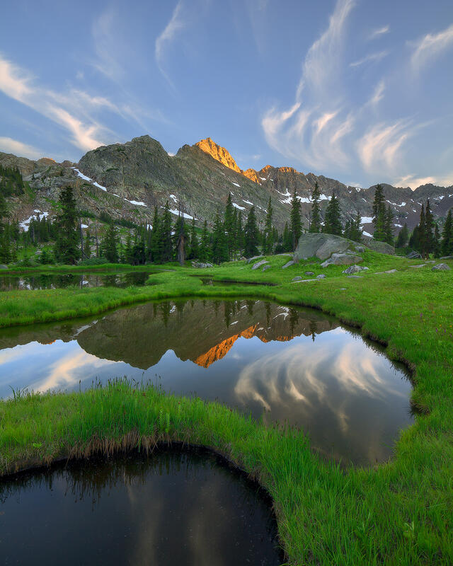 Green grass and winding, still water lead into the scene with the pine trees, wispy white clouds are reflected on the water; a red mountain in the background.