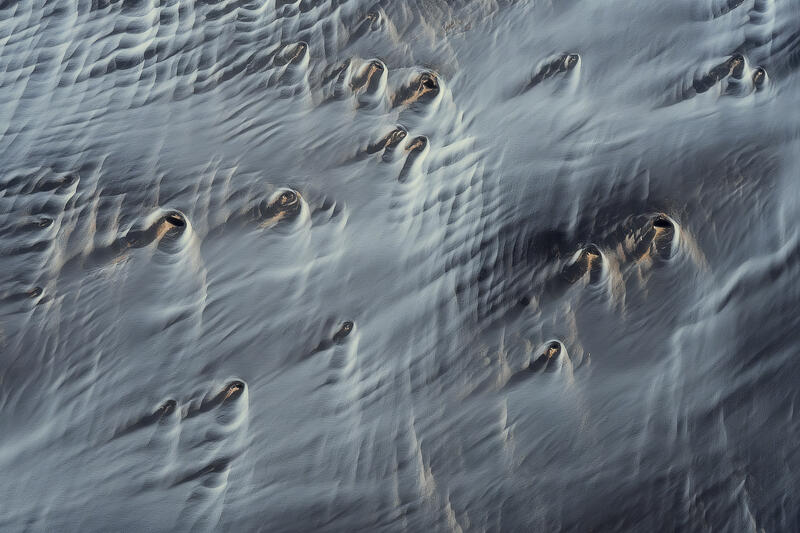 Ghostlike volcanic formations emerge from flowing ribbons of wind-shaped sediment across Iceland's stark volcanic plains.