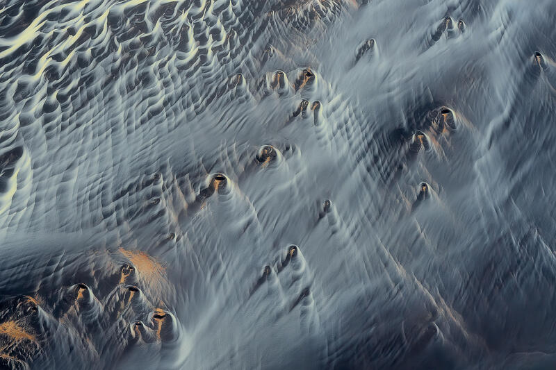 Eroded volcanic formations emerge like silent figures through flowing glacial sands in Iceland.
