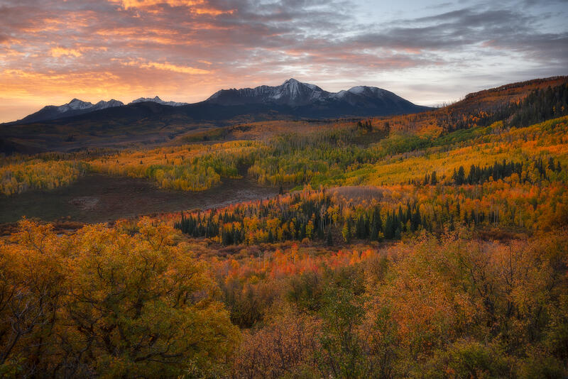 Layers of brilliant Colorado fall color sweep toward snow-dusted mountains beneath a glowing autumn sunset.