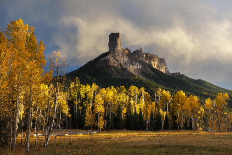 Mountain peak stands tall with clouds behind it and yellow aspen trees shining in the sunlight below.