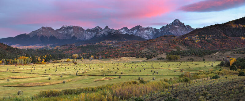 A mountain range sits behind a field full of hay bales while the sun sets and colors the sky pink.