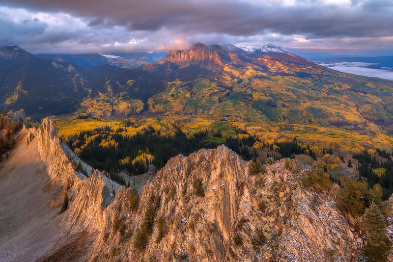 Jagged peaks lead into the image with a valley of yellow and green aspen trees and mountains in the distance are covered with some clouds and snow at sunrise.