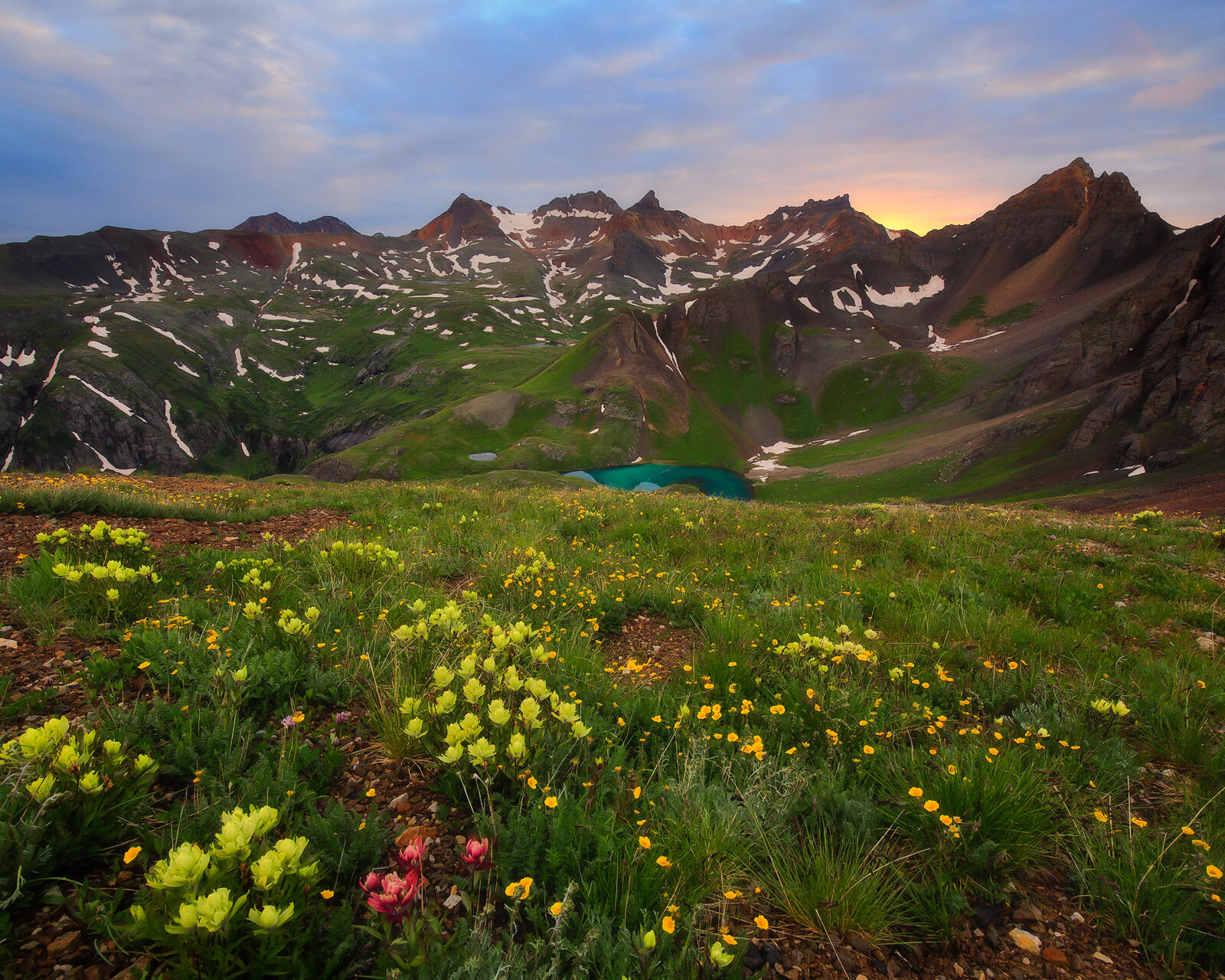 Alpine lake is seen below with a field of springtime wildflowers in front and a range of mountains in on the horizon as the sunsets.