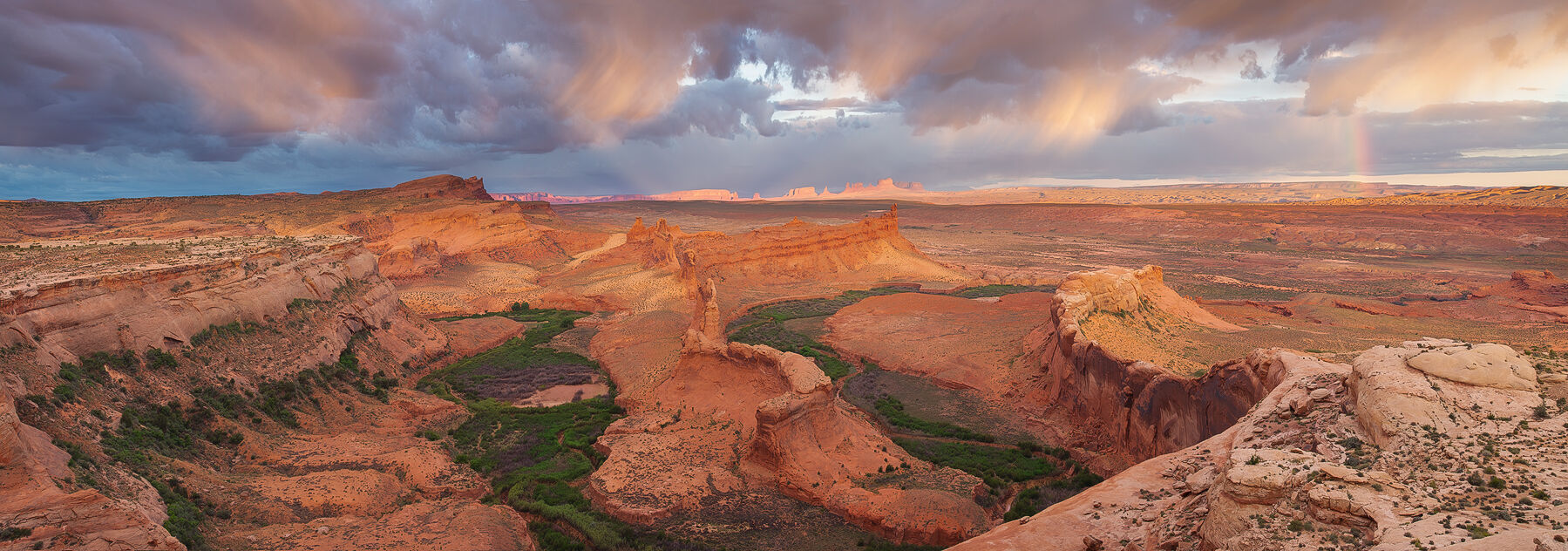 Ridges stand above the lush green floor of this desert scene clearing storm clouds in the distance and and a rainbow as the sun peers through the clouds.