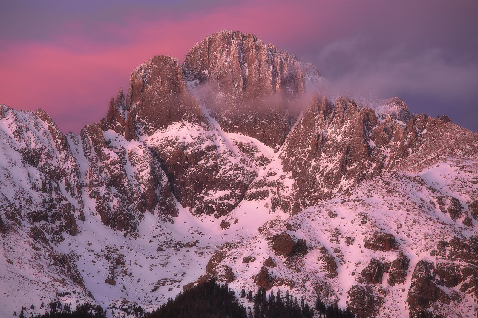 The top of Crestone Peak is seen slightly shrouded in clouds with pink skies at sunrise. Snow on the mountains glows with the sun.