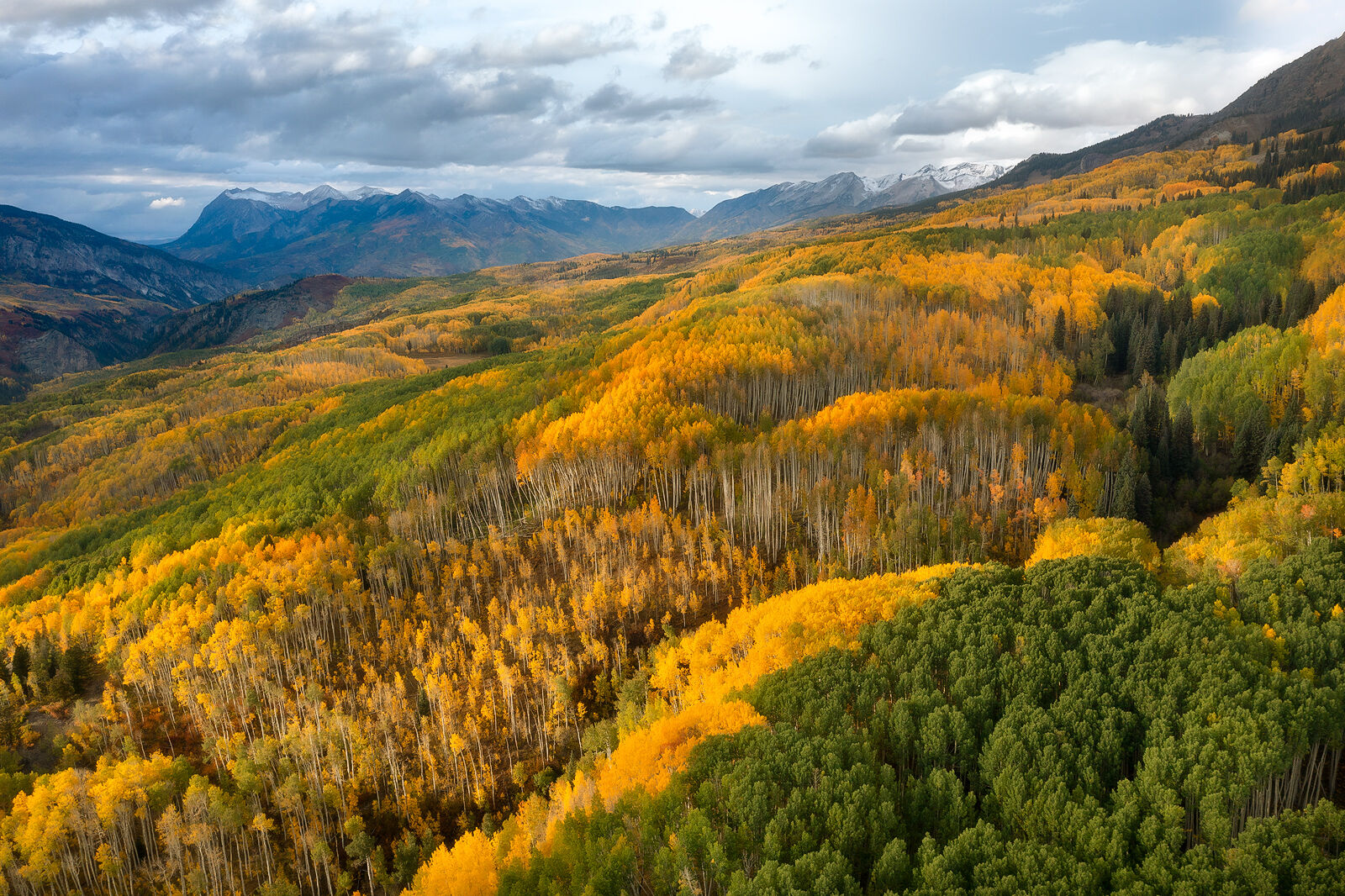 Raggeds Sunset Mountains in the distance sit just beneath the clouds as the valley below is filled with yellow, orange and green aspens.