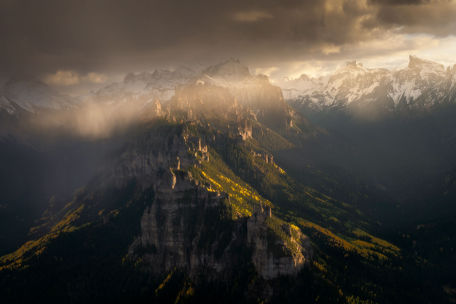 A mountain ridge is hit by sunlight as the sun sets and clouds obscure the distant surrounding mountains dusted in snow with a golden aspen tree hillside on the