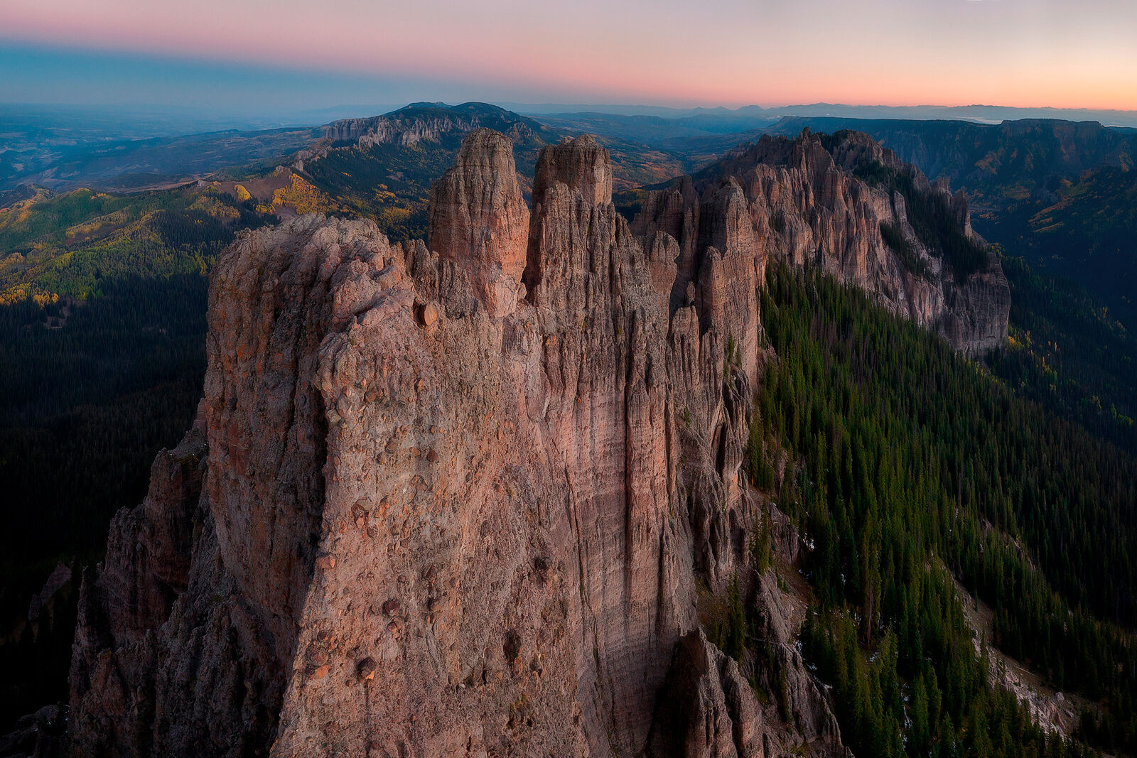 A mountain ridge is seen up close at sunrise with a pink and orange sky and other ridges, fall color aspen trees, and spruce trees in the valley.