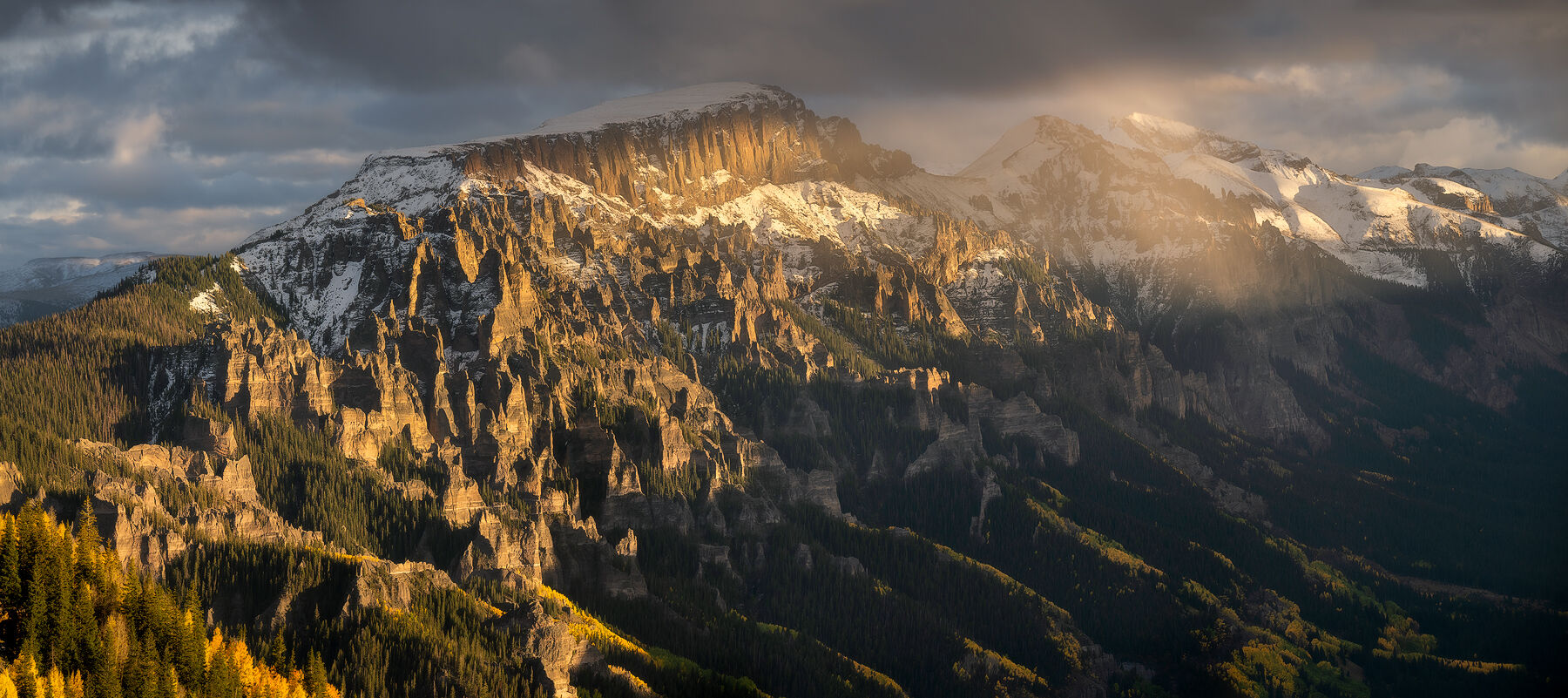 A moody panoramic scene of a mountain skyline clouds making way for the sun to hit the tops of the mountains and yellow-leaved aspen trees and spruce trees in t