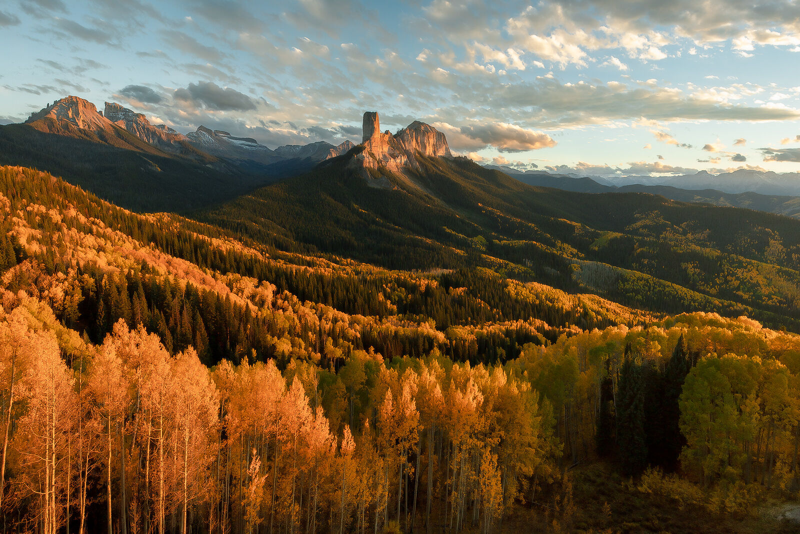 A tall, columnar rock stands in the background with bright yellow aspen trees shining their leaves as the sun lights up the scene from the side as it sets.