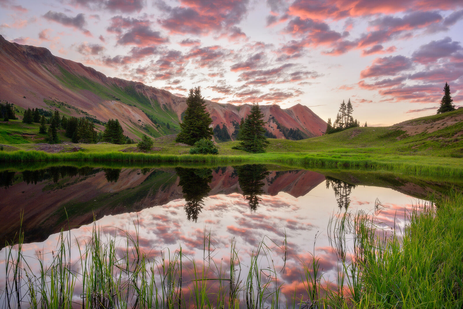 Green grass surrounds a still lake with mountains in the background and pink clouds in the sky at sunset - all of which is reflected on the water.