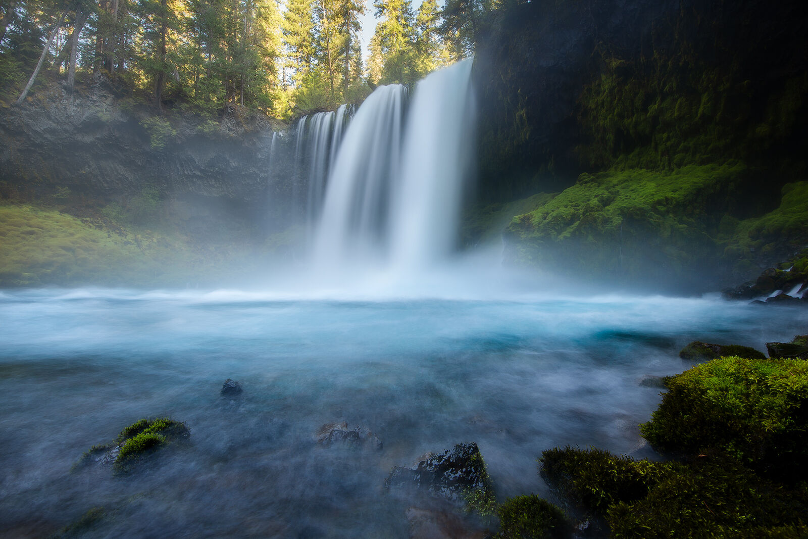 A waterfall in the forest plunges into a deep blue pool of water and the sun highlights the blue color of the water.