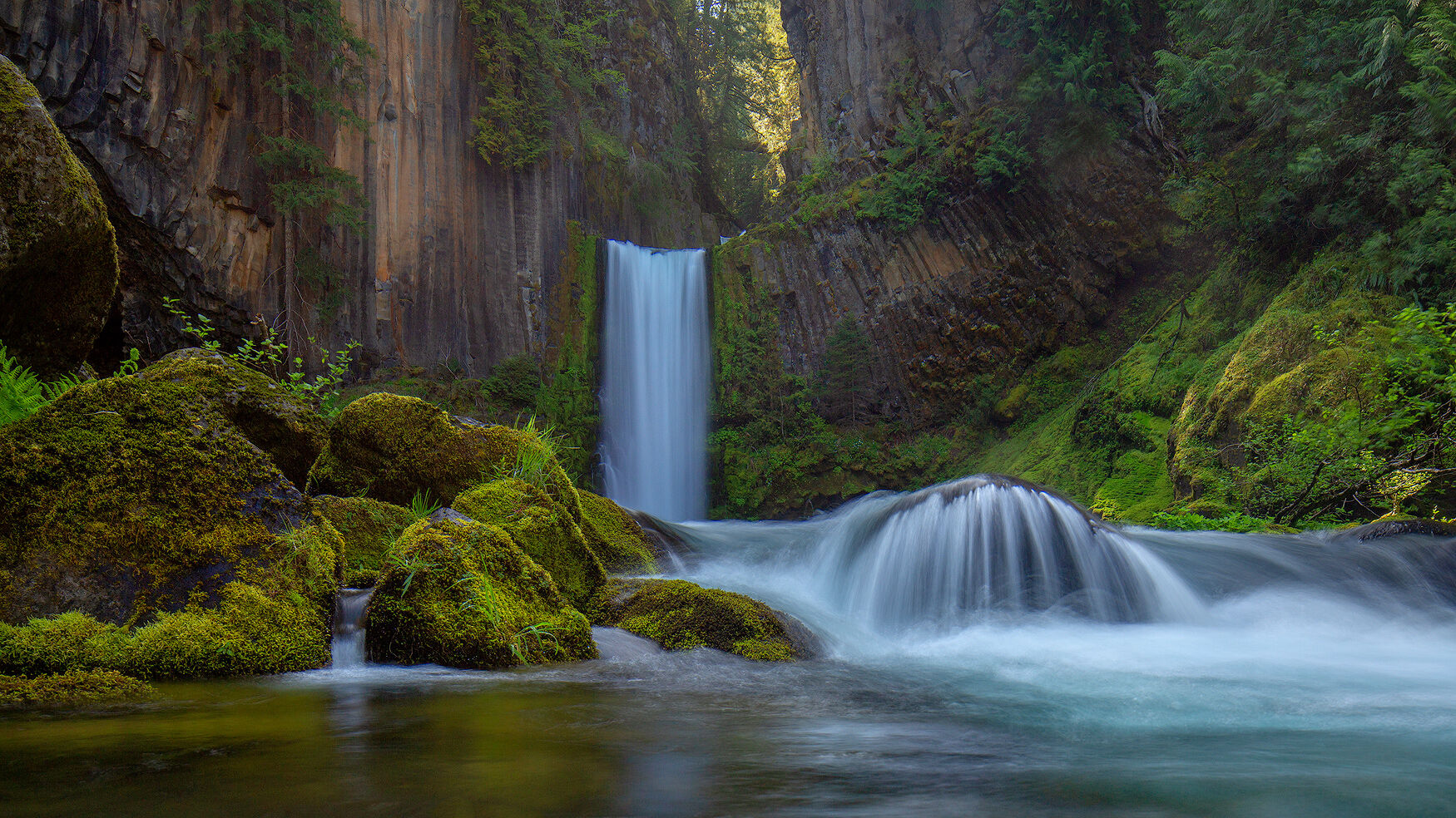 Oregon's Secrets A two-layer waterfall with vertically gouged rock stretching up and lush green foliage and mossy covered rocks meeting the pool of water.