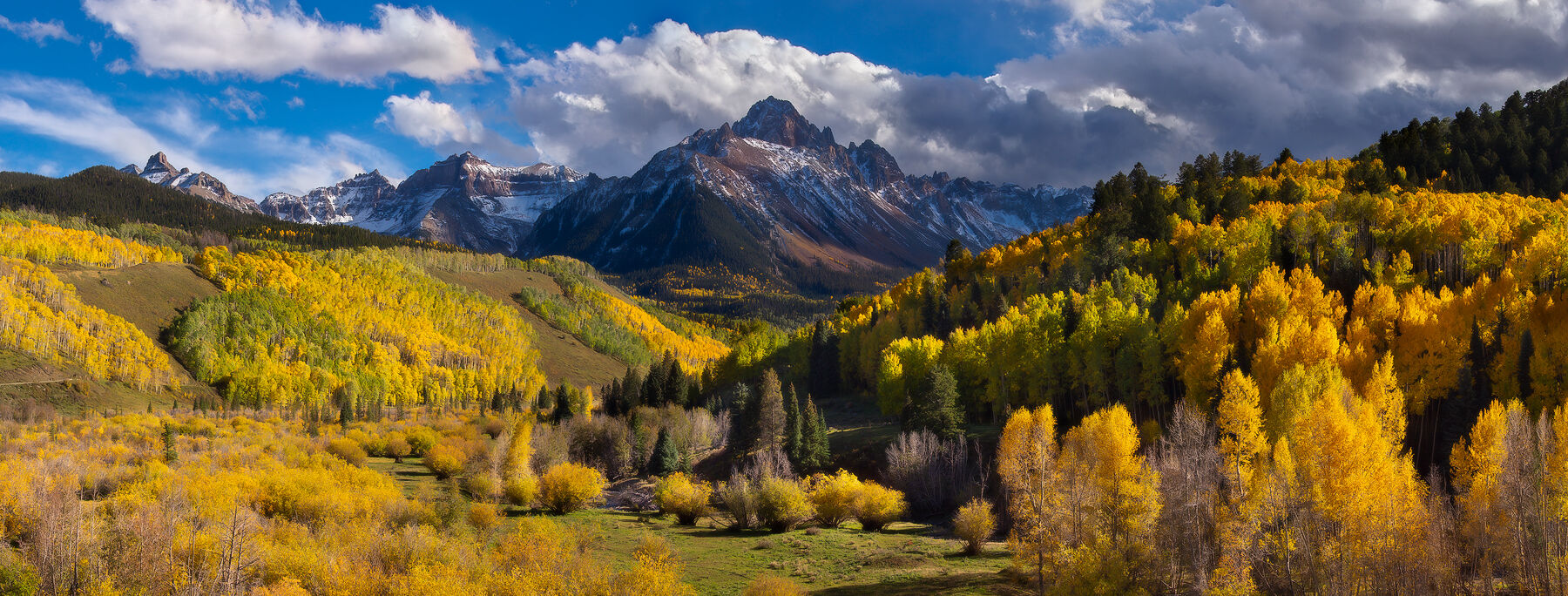 Mountains in clouds in the distance and yellow trees throughout the valley are lit up by the sunlight shining through the clouds.