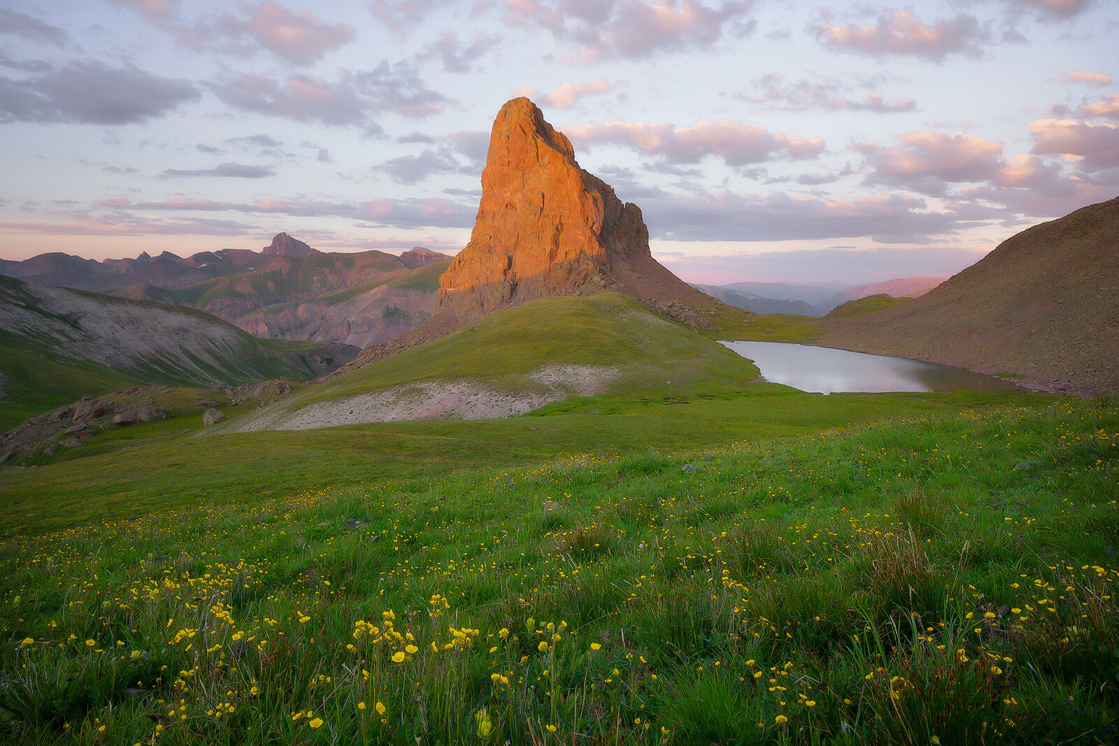 Mountain peak stands solo surrounded by a meadow of green grass and a mountain range in the background and a small lake. All while the sun sets a pink sky