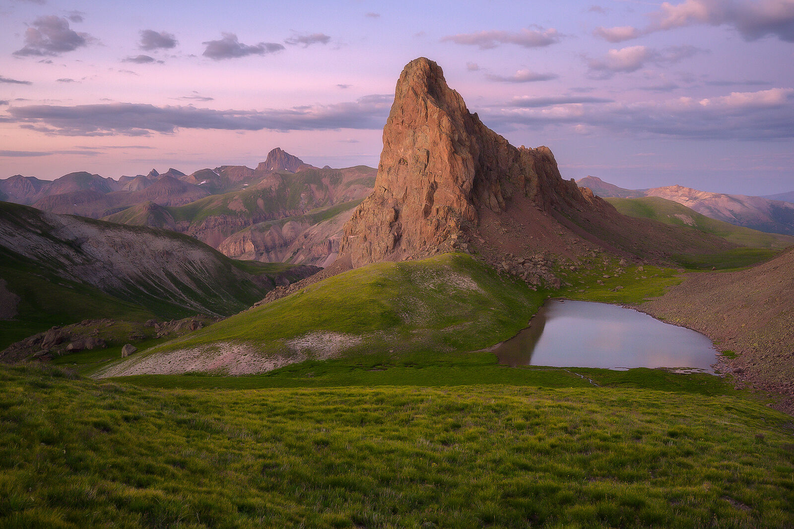 Prominent mountain peak stands with green grass surround it at sunset with pink clouds in the sky and a horizon full of mountain peaks.