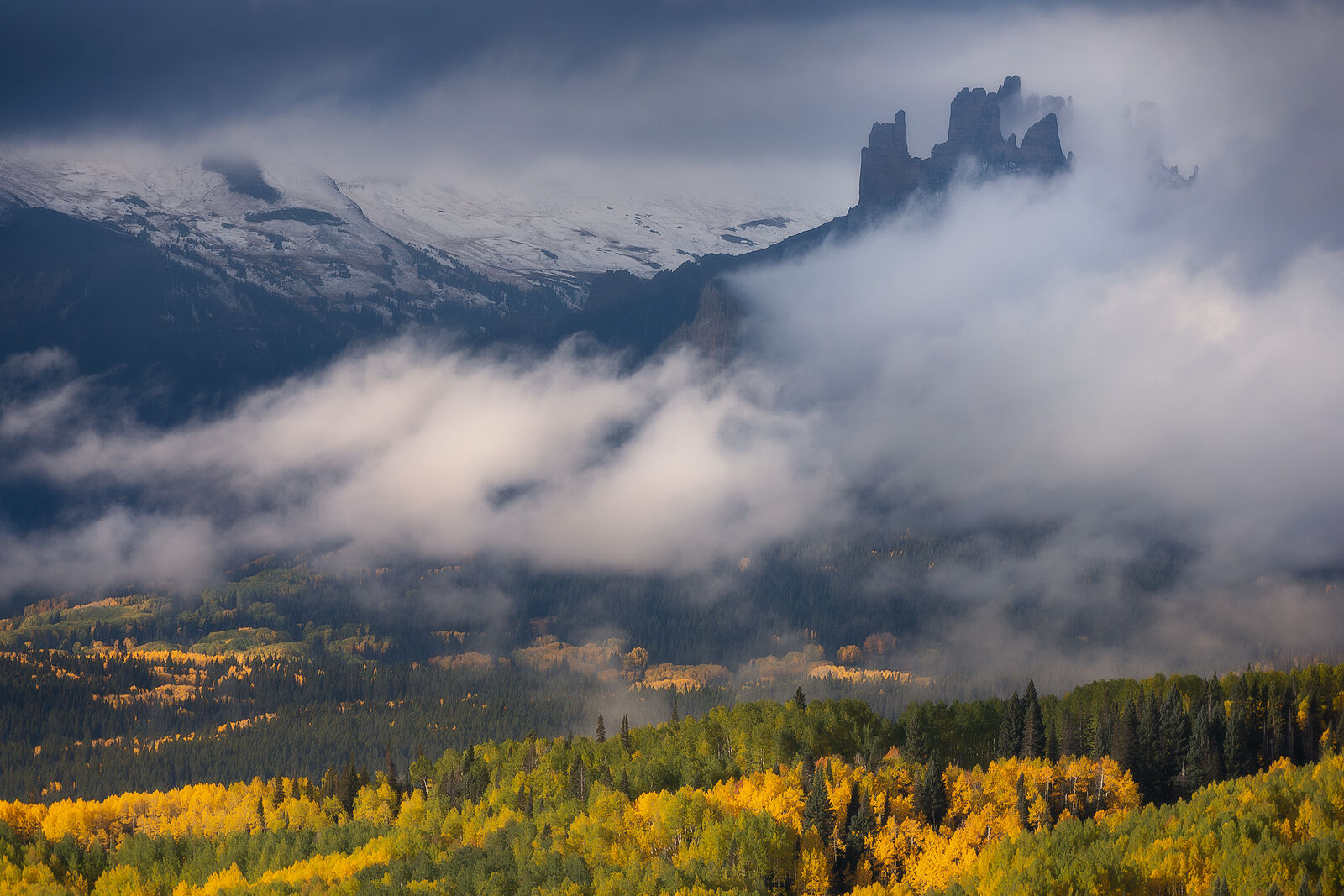 Dramatic clouds hide rocks that look like castle spires in the background with snow covered mountain peaks and yellow aspens in front.