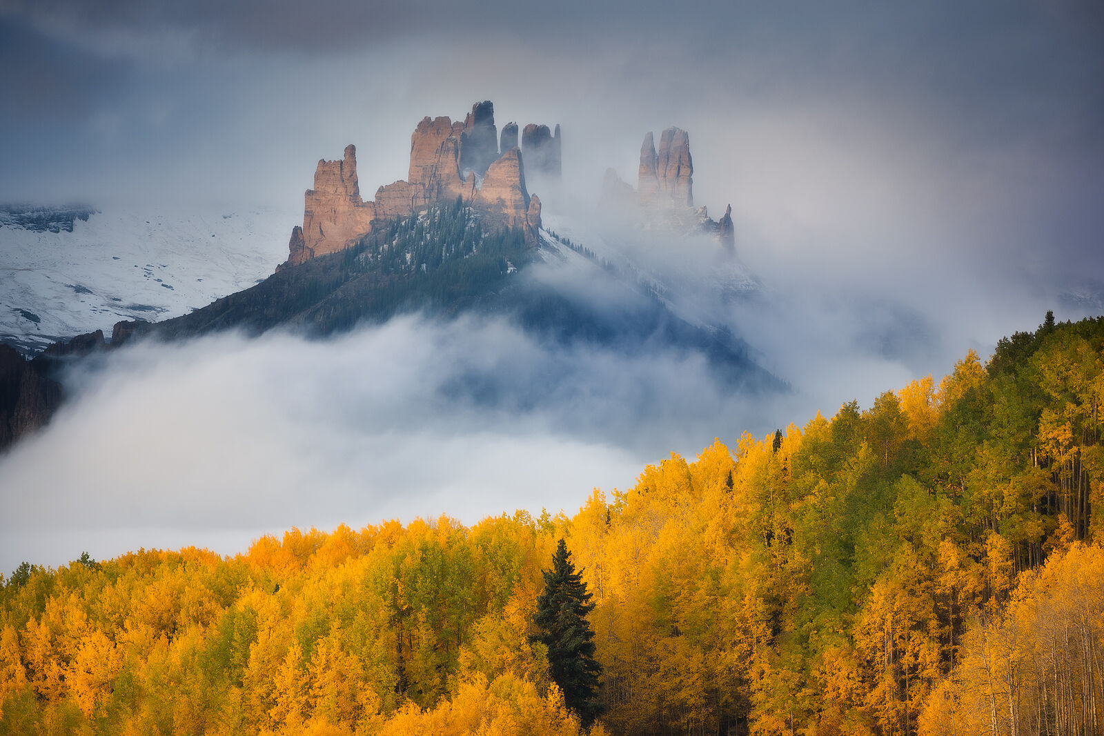 Breaking Through Mountains shaped like castle spires show through and above clouds as the sun breaks through and bright yellow aspens frame the bottom corner of the image.