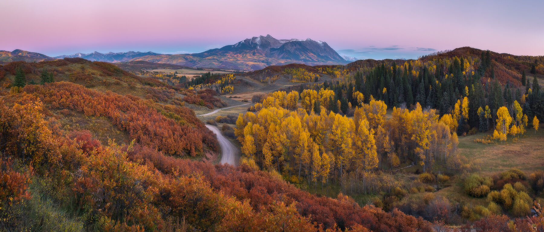 Fall color hillsides are seen with a dirt road winding through the scene and mountains in the background.