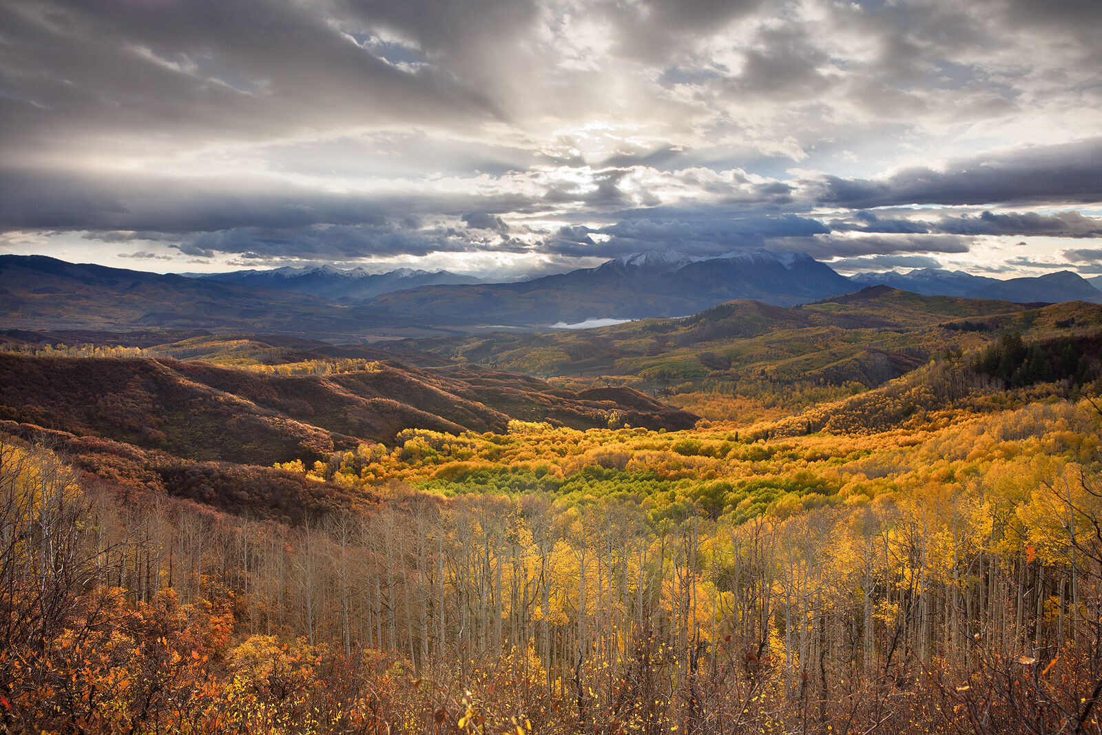Mountains on the horizon have peaks nearly hidden by clouds and the valley below is full of yellow and orange aspens and some without leaves.